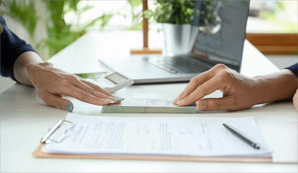 A close-up image of two people sitting at a desk, discussing "Security Deposit Alternatives." One person, with turquoise nail polish, is handing over a stack of cash to another person. In the foreground, there is a clipboard with a document and a pen resting on it. In the background, a laptop and a potted plant can be seen, suggesting a professional office environment. The image highlights the concept of alternative options for security deposits in rental agreements.