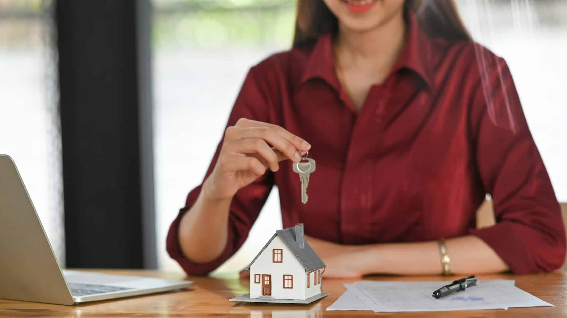 Person in a red shirt holds a key over a small model house on a desk with a laptop and papers nearby, suggesting a home purchase or rental scenario.